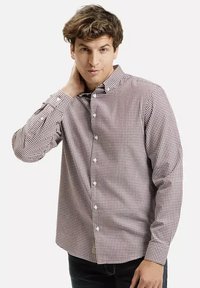 Mężczyzna w czerwono-białą kratkę koszuli zapinanej na guziki, dotykający szyi lewą ręką, patrzący w kamerę na tle białym.