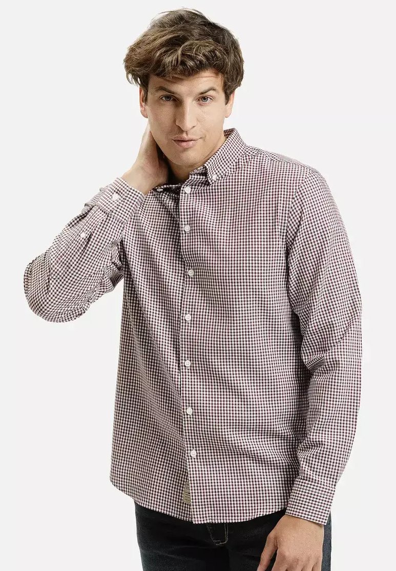 Mężczyzna w czerwono-białą kratkę koszuli zapinanej na guziki, dotykający szyi lewą ręką, patrzący w kamerę na tle białym.