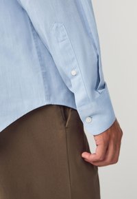 Camicia azzurro chiaro a maniche lunghe con vestibilità sartoriale, polsini con bottoni e tessuto dalla trama liscia, abbinata a pantaloni marroni.