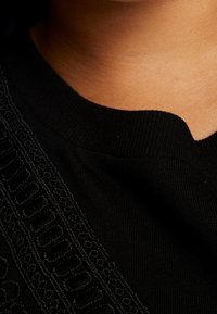 Czarna bluzka z fakturowanym, haftowanym detalem wzdłuż linii dekoltu, z zaokrąglonym kołnierzykiem i miękką teksturą tkaniny.