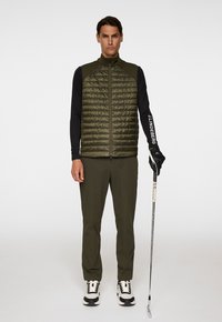 Gilet trapuntato verde oliva con colletto alto sopra una maglietta nera a maniche lunghe, abbinato a pantaloni verde oliva e sneaker nere, mentre tiene un bastone da golf.