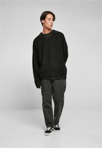 Suéter negro oversized con un profundo escote en V y un patrón de punto texturizado. Lleva una camisa negra por debajo y se combina con jeans sueltos de un gris oscuro y zapatillas negras.