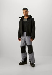 Črna vodoodporna jakna čez sivo majico z dolgimi rokavi, kombinirano s sivimi hlačami s črnimi madeži. Model stoji na nevtralni podlagi.