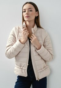 Jasno beżowa kurtka puchowa z pionowym pikowaniem, wysokim kołnierzem i zapięciem na zamek, z elastycznymi mankietami i pasem.