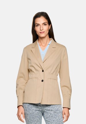 Frau in einer beige geknöpften Jacke über einem hellblauen Oberteil und gemusterten Hosen, die vor einem schlichten Hintergrund steht, nach vorne blickt und einen entspannten Gesichtsausdruck hat.