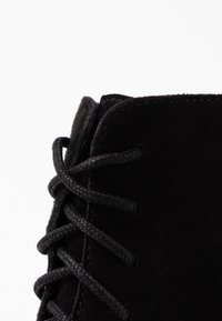 Černé semišové kotníkové boty s černými tkaničkami provlečenými zesílenými očka, které vykazují měkkou strukturu a zaoblený tvar nahoře.