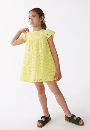 Vestido de algodón amarillo con mangas cortas, estampado de ojal y cintura fruncida. Combinado con sandalias oscuras que tienen hebilla y suela texturizada.