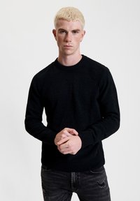Czarny sweter robiony na drutach z okrągłym dekoltem, ściągaczami na rękawach i u dołu. Nosi go osoba z krótkimi, jasnymi blond włosami i neutralnym wyrazem twarzy.