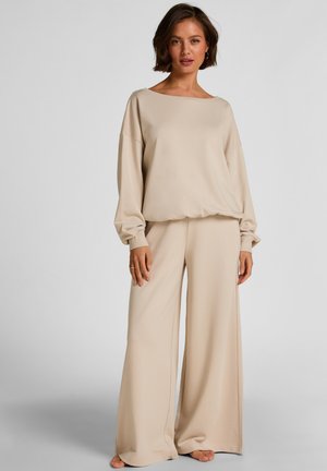 Vrouw staat blootsvoets, draagt een beige loszittende top met lange mouwen en wijde broekspijpen tegen een effen lichte achtergrond.