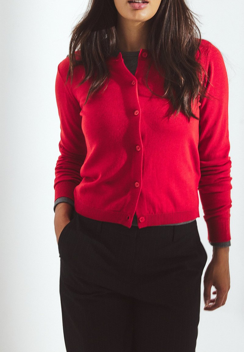 Red cardigan with button closure, ribbed cuffs and hem, worn over a gray shirt. Paired with black striped trousers. Smooth texture.