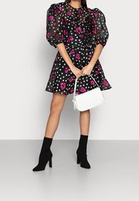 Robe noire à manches bouffantes ornée de motifs floraux roses et de pois blancs, assortie à un sac à main en osier blanc et des bottines à talons noires.