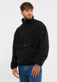 Pullover nera in pile con colletto alto, zip frontale a metà e tasca anteriore. Texture morbida e vestibilità comoda. Indossata sopra jeans blu.