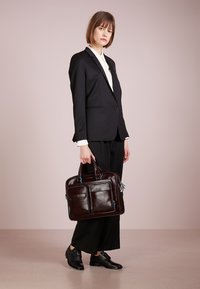Valigia in pelle marrone con due tasche frontali, dettagli in cerniera blu e doppi manici. Il modello indossa un completo nero e scarpe nere.