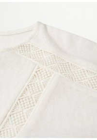 Camiseta de algodón blanco con bordado texturizado y patrones geométricos a lo largo de la costura del hombro. Tejido suave con un acabado blando.