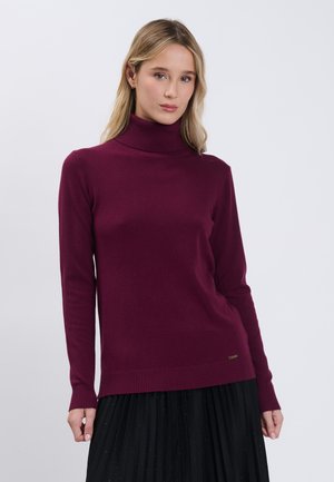 Ung kvinde iført en langærmet bordeaux rullekravesweater og et sort plisseret skørt, stående mod en ensfarvet lys baggrund.