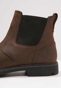 Botte Chelsea en cuir marron avec un panneau élastique noir, un bout arrondi et une semelle en caoutchouc texturée avec un logo embossé sur le côté.