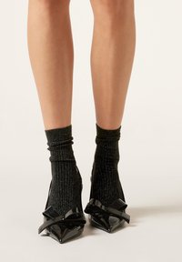 Sorte laklæderstilethæle med spids tå og skinnende sløjfer, kombineret med sorte ribbede metallicstrømper, der fremviser en struktureret, glitrende overflade.