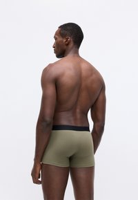 Bóxer verde oliva con una cinturilla negra, hecho de una tela suave, mostrado en un hombre de espaldas, destacando un diseño simple y ajustado.