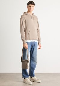 Jonge man in een beige hoodie, blauwe jeans en beige sneakers, met een taupekleurige schoudertas met het BOSS-logo, staand op een effen achtergrond.