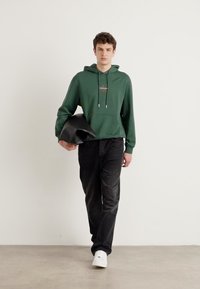 Felpa verde con cappuccio e lacci, jeans neri e scarpe bianche. Porta una borsa nera. Outfit casual con una vestibilità rilassata e texture morbide.