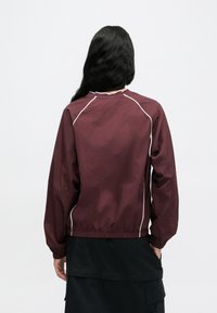 Bordová ľahká bunda so zaobleným výstrihom, elastickými manžetami a elastickým pásom. Biele lemovanie pozdĺž švov zvýrazňuje jej dizajn.