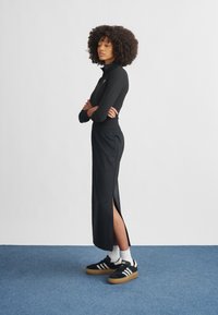 Top preto justo com fecho de correr, combinado com uma saia maxi preta com fendas laterais. Usado com sapatos escuros e meias brancas, num fundo azul.