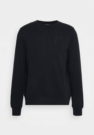 Melnā sweatshirt, kas izgatavota no mīkstas auduma, ar apaļu kaklu un garām piedurknēm. Uz priekšu ir redzams teksturēts K logo kreisajā krūtī.