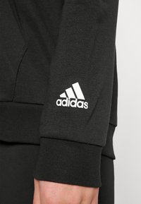 Sweat-shirt noir à poignets côtelés ; comporte un logo adidas blanc sur la manche. Le matériau semble doux et texturé.