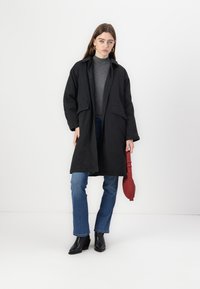 Black long coat with a collar, two front pockets, worn over a gray turtleneck. Paired with blue jeans and black ankle boots. Red bag held in hand.