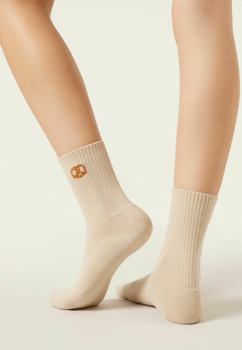 Cream ribbed socks worn on feet, one foot raised showing brown pretzel embroidery on ankle.