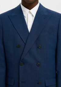 Blazer doppiopetto blu navy realizzato in tessuto testurizzato, con bottoni neri, rever a scollo e una tasca sul petto sopra una camicia bianca.