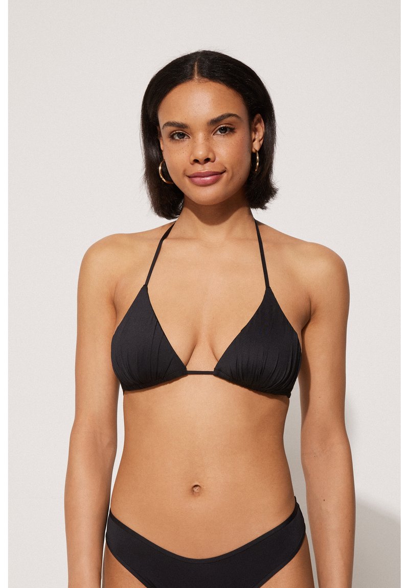 Femme aux cheveux bruns mi-longs portant un haut de bikini noir style halter et un bas de bikini noir assorti sur un fond uni.