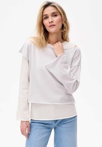 Helles graues Langarmoberteil mit einem geschichteten Design, strukturiertem Stoff und offenen Schulterdetails. Kombiniert mit hellblauen Jeans.