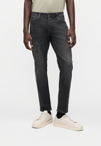 Σκούρο γκρι τζιν παντελόνι από τζιν ύφασμα με ίσια γραμμή, πεντατσέπη σχέδιο και θηλιές για ζώνη, συνδυασμένο με ανοιχτόχρωμα αθλητικά παπούτσια και μαύρες κάλτσες.