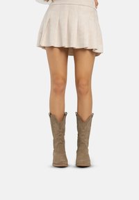 Botas de vaquero de ante marrón con patrones florales bordados, punteras puntiagudas y lengüetas de tirón. Llevadas con una falda plisada ligera.