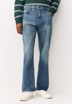 Personne portant un jean bootcut bleu délavé, un t-shirt vert à rayures manches longues et des baskets blanches, debout avec une main dans la poche.
