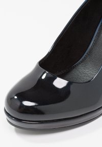 Décolleté in vernice nera con punta arrotondata, esterno liscio e lucido, e una piattaforma bassa. L'interno presenta un rivestimento morbido e testurizzato.