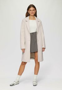 Lichtbeige lange jas met notch-lapels, gedragen over een witte top en een grijs pinstriped rok. Het model draagt zilveren platte schoenen en witte geribde sokken.