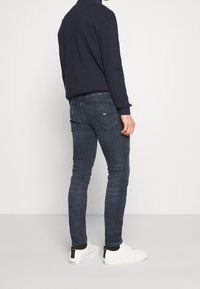 Jersey azul marino, jeans ajustados azul oscuro y zapatillas deportivas blancas; el denim muestra un ligero desgaste, diseño minimalista y bolsillos traseros visibles con el logo.