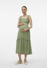 Vestido de maternidad verde hecho de una tela ligera con un cuerpo fruncido, tirantes con volantes y falda en capas, combinado con zapatos planos beige.