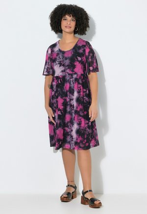 Femme aux cheveux bouclés portant une robe tie-dye rose et noire à manches courtes jusqu'aux genoux et des sandales à plateforme noires, debout sur fond blanc.