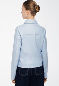 Lyseblå læderjakke med krave, tætsiddende design, lange ærmer og detaljer på bagsømmen, parret med mørke denimbukser.