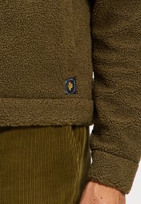 Oliivroheline fleece-pullover, millel on tekstuuriga pind ja soonikvarrukad. Üksikasjadena on allservas õmmeldud brändi silt.