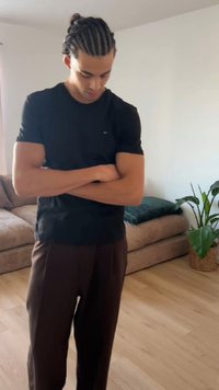 Νέος άνδρας με πλεξούδες, φορώντας μαύρο πουκάμισο και καφέ παντελόνι, στέκεται με σταυρωμένα χέρια και κοιτάζει προς τα κάτω σε ένα σαλόνι.