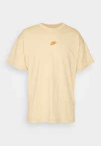 Beige kortärmad Nike T-shirt med liten orange Nike-logotyp centrerad på bröstet, visad på en vit bakgrund.