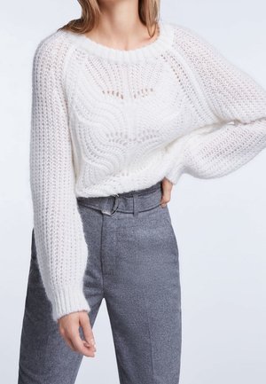 Femme portant un pull tricoté blanc avec des motifs texturés et un pantalon taille haute gris clair avec un détail ceinture.