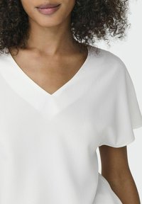 Top blanc avec un décolleté en V et des manches courtes, fabriqué en tissu texturé avec une finition douce. Détails de couture le long du col et des manches.