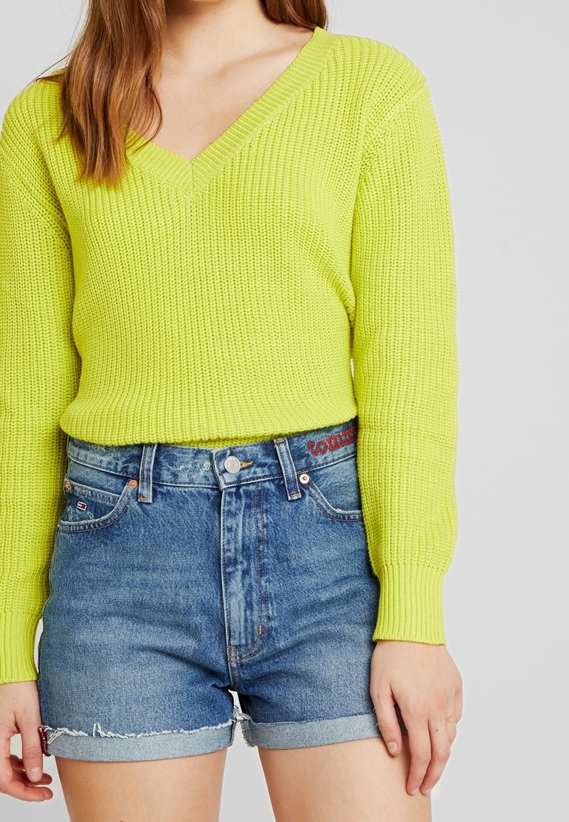Pull en maille jaune vif à col en V, associé à un short en denim bleu avec un ourlet effiloché et un discret détail de marque sur la ceinture.