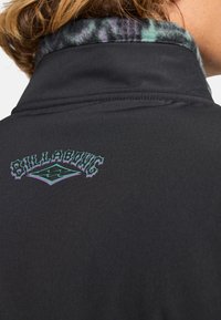 Veste noire avec un col haut dotée d'une doublure en polaire douce et à motifs. Le dos présente un logo "BILLABONG" coloré dans une police stylisée.