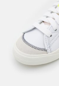 Witte sneaker met een suède neuskap, zwarte stiksels en een gestructureerde rubberen zool. Bevat witte veters en minimalistische designelementen.
